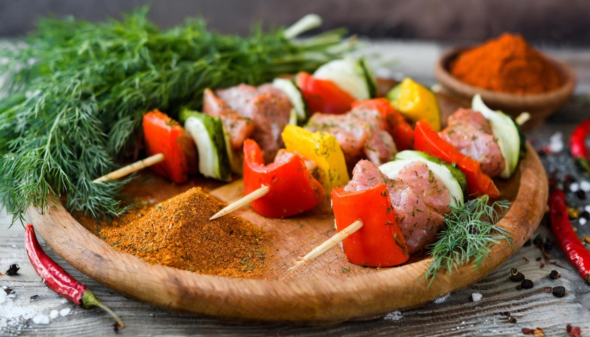 Des brochettes de viande et de legumes sont posées sur un plateau en bois. Il y aussi de l'aneth et du paprika