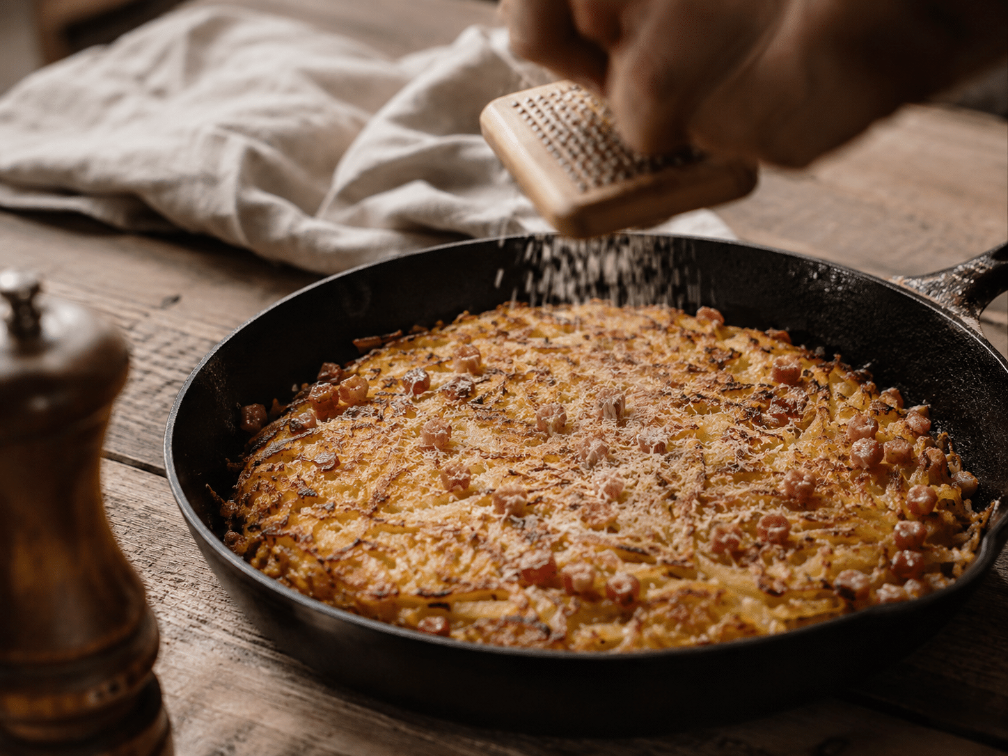 Une poêle en fonte avec le rösti bernois et quelqu'un qui râpe la muscade.